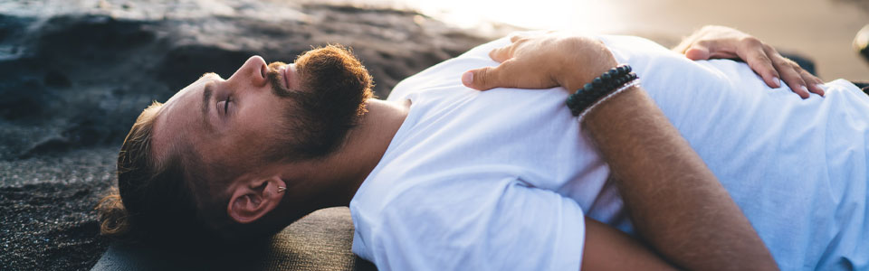 Mann entspannt am Strand in Meditation Ein Mann mit Bart liegt entspannt auf einer Yogamatte am Strand.