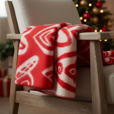 Rote Decke auf Stuhl neben Weihnachtsbaum Nahaufnahme einer roten Decke mit weißen Mustern, die über einer Armlehne eines Holzstuhls hängt, im Hintergrund ein Weihnachtsbaum mit Lichtern und Dekorationen.