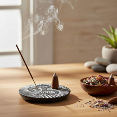 Räucherstäbchen mit Rauch auf einem Tisch Ein brennendes Räucherstäbchen, das auf einem dekorativen Ständer mit Rauch auf einem Tisch positioniert ist, daneben ein Gefäß mit getrockneten Blüten und zenartigen Steinen im Hintergrund.