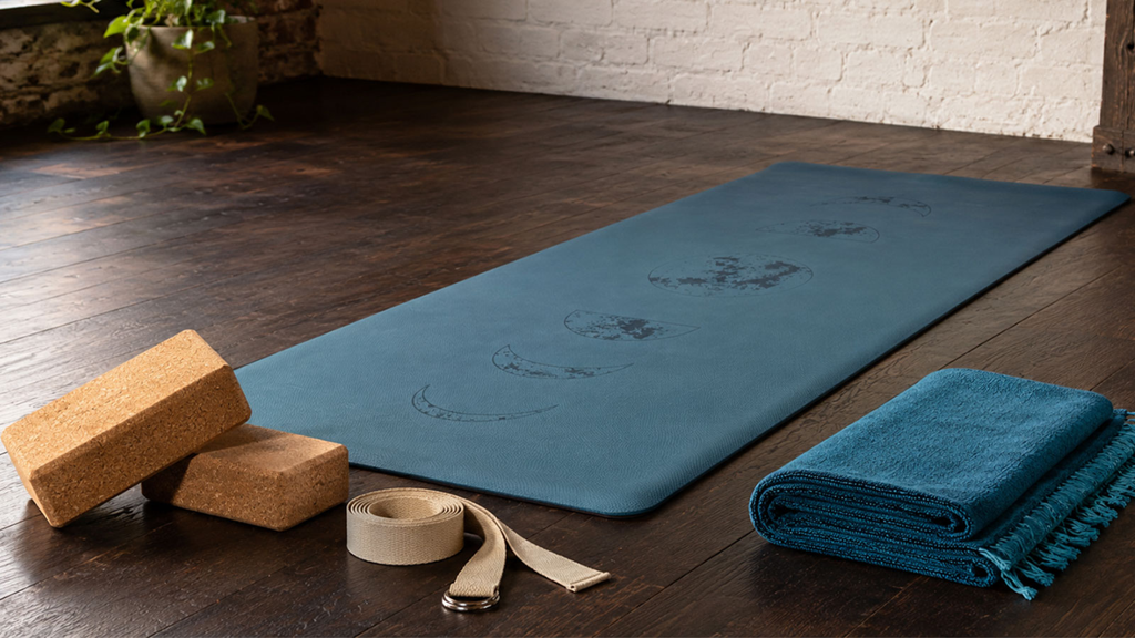 Yoga-Zubehör auf Holzboden Auf einem dunklen Holzboden liegen eine blaue Yogamatte mit Mondphasenmuster, zwei Korkblöcke, ein Yogagurt und eine gefaltete blaue Decke.