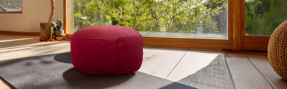 Ein rotes Meditationskissen auf einem Boden nahe einem großen Fenster, im Hintergrund sind grüne Bäume zu sehen.