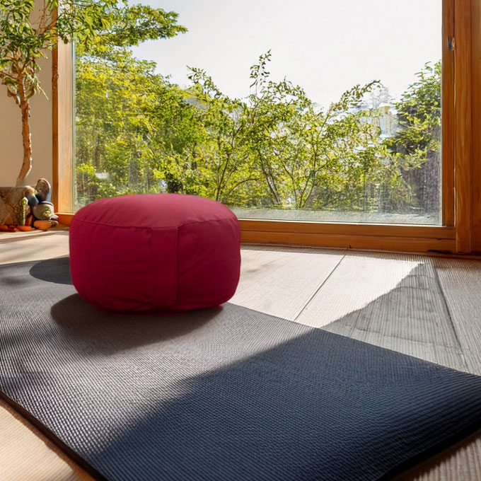 Ein rotes Meditationskissen auf einer Yoga-Matte vor einem großen Fenster mit Blick auf grüne Bäume.