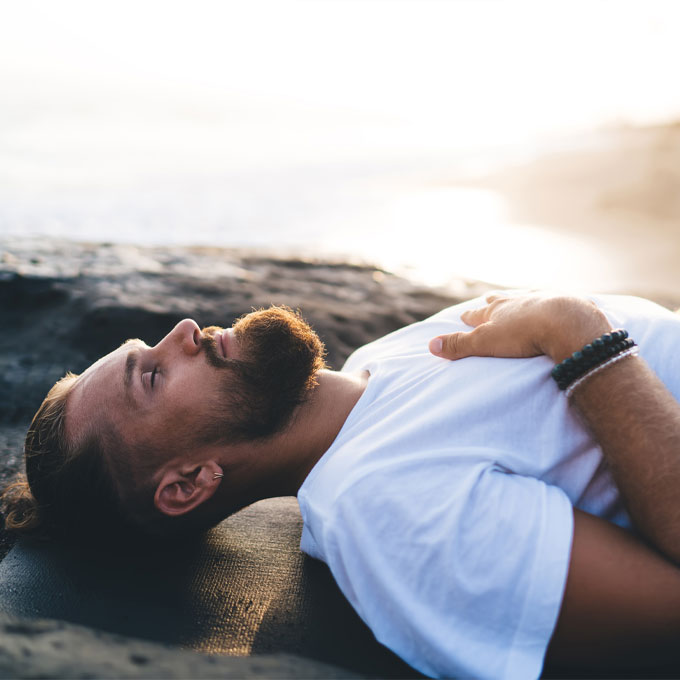 Mann mit Bart, der am Strand bei Sonnenuntergang auf einer Matte liegt und entspannt.