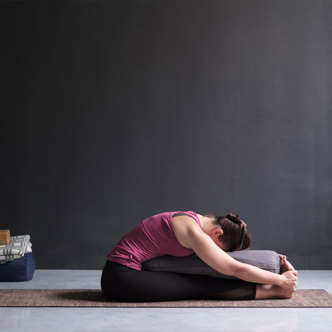 Eine Frau in Leggings und Tanktop macht eine Yogaübung in einem dunklen Raum. Sie sitzt auf einer Matte und beugt sich mit einem Kissen unter dem Oberkörper nach vorne.