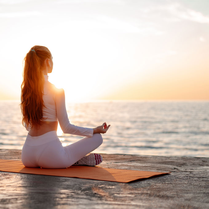 Eine Person meditiert im Lotussitz auf einer Yogamatte am Meer bei Sonnenuntergang.