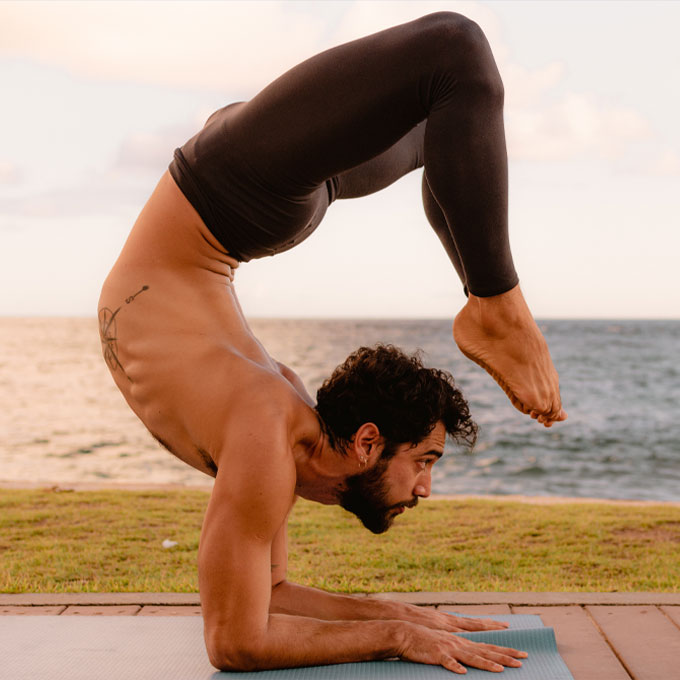 Ein Mann in einer fortgeschrittenen Yoga-Pose auf einer Matte am Strand, die Sonne scheint im Hintergrund.