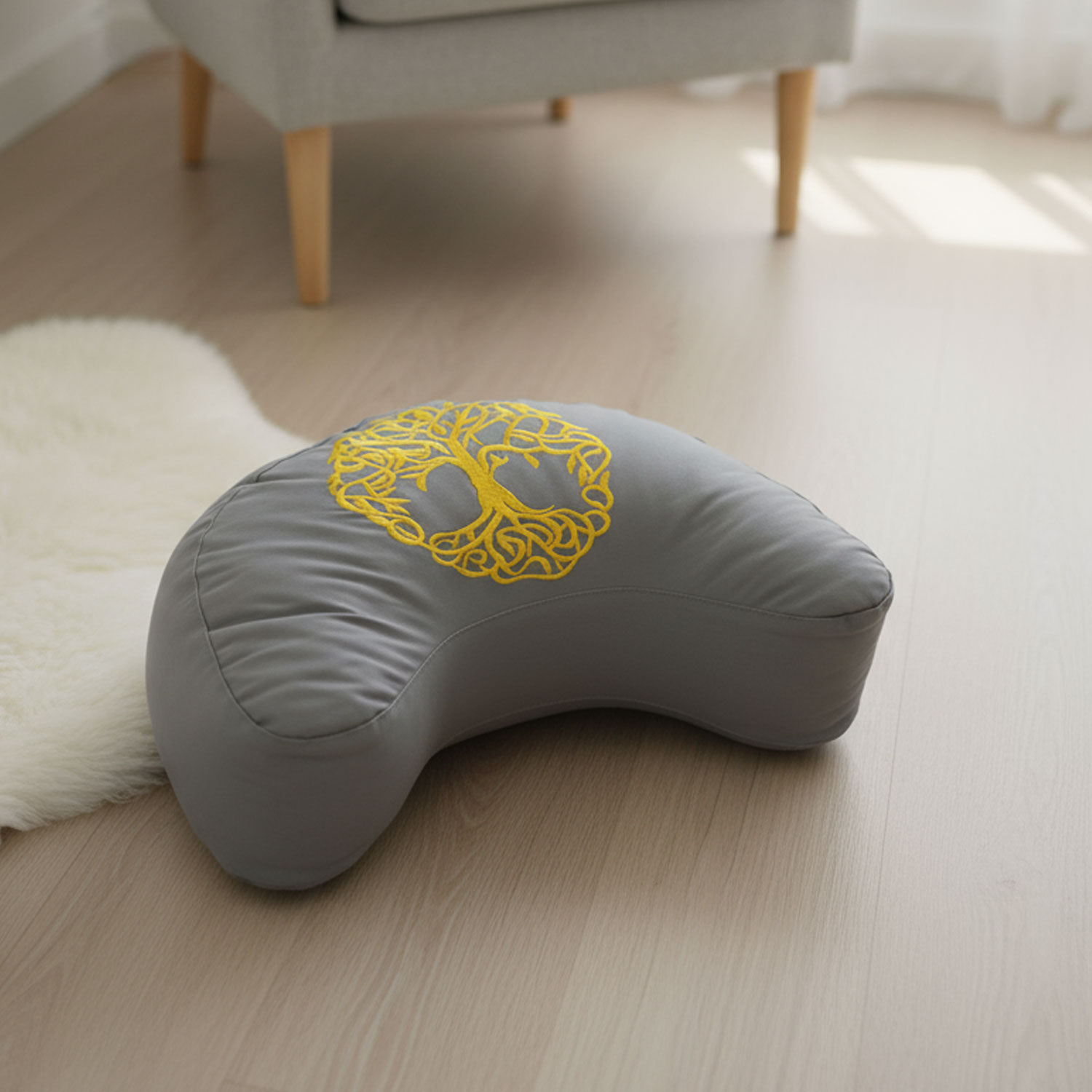 Graues, mondförmiges Meditationskissen mit gelbem Baumdesign auf hellem Holzboden neben einem weißen Teppich.