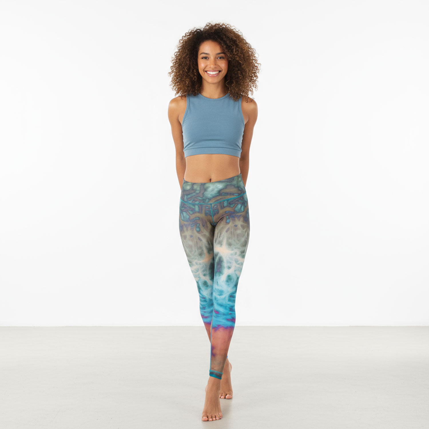 Junge Frau mit lockigen Haaren in bunter Sportleggings und blauem Top steht barfuß und lächelt