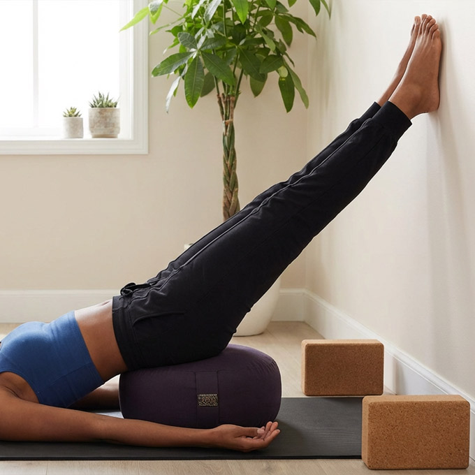Eine Frau macht Yoga-Übung, liegt auf einer Matte mit den Beinen an der Wand, gestützt von einem Kissen.