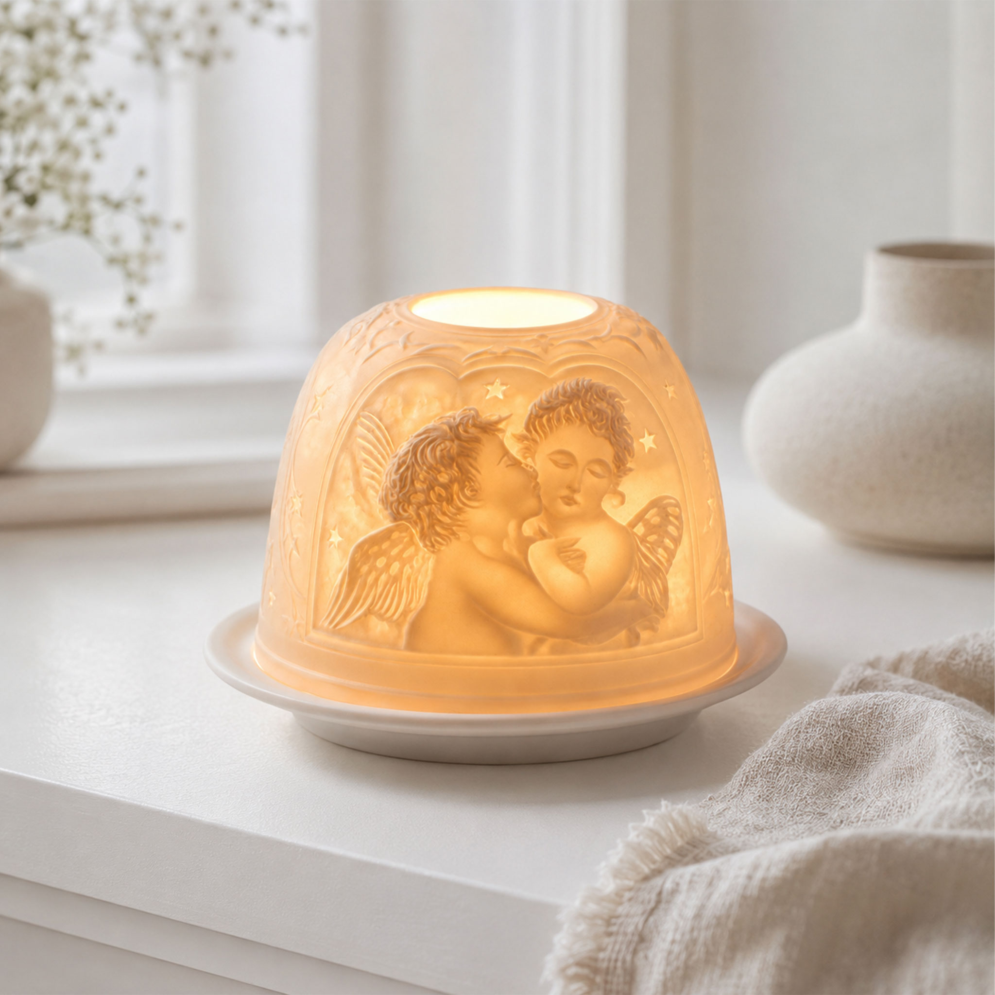 Eine warme, beleuchtete Porzellanlampe mit eingravierten Engelfiguren auf einem weißen Unterteller in einer hellen Wohnumgebung.