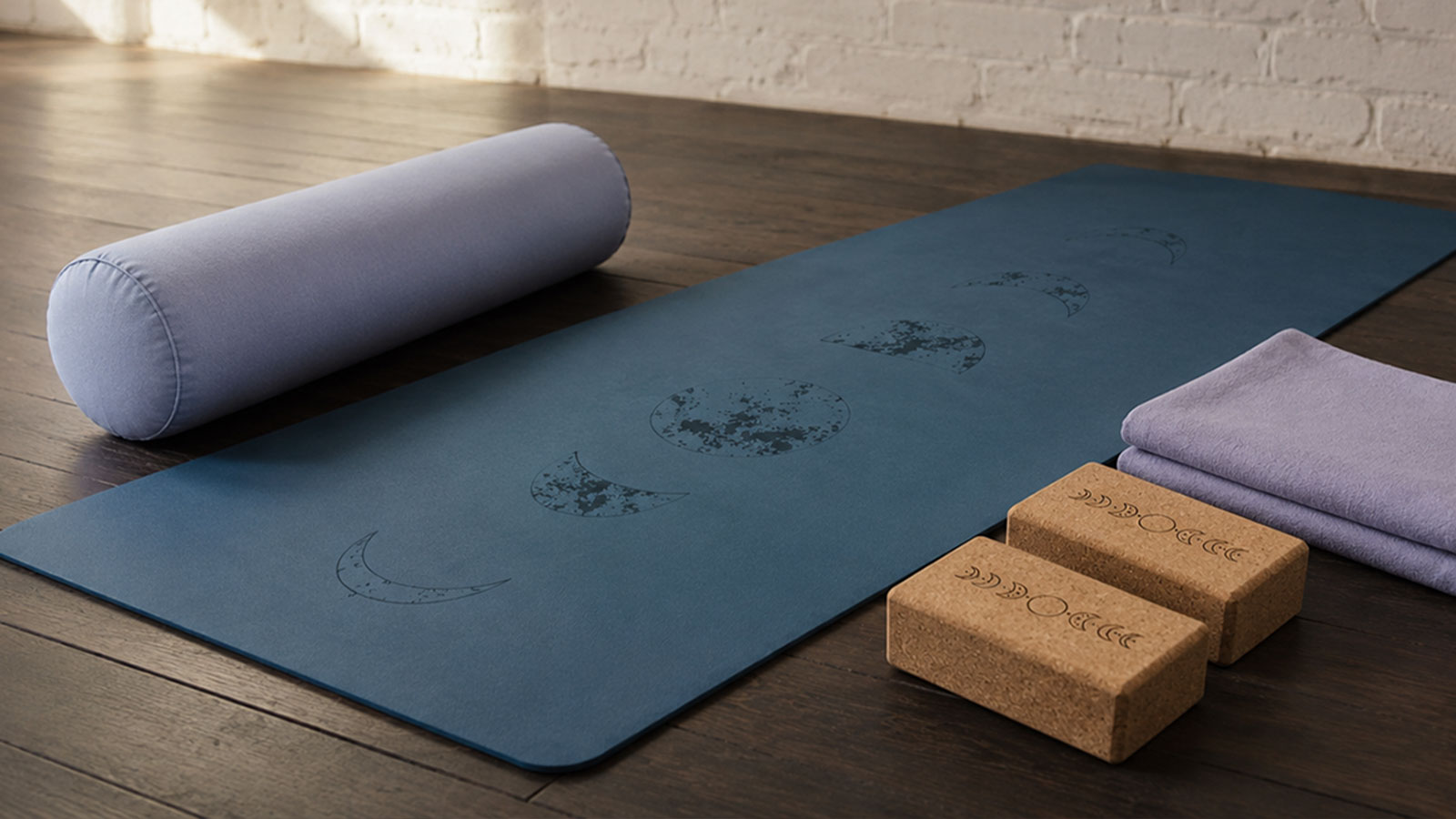 Eine blaue Yoga-Matte mit Mondphasen-Motiv, daneben ein runder Bolster, zwei Korkblöcke und gefaltete Tücher auf einem dunklen Holzboden.