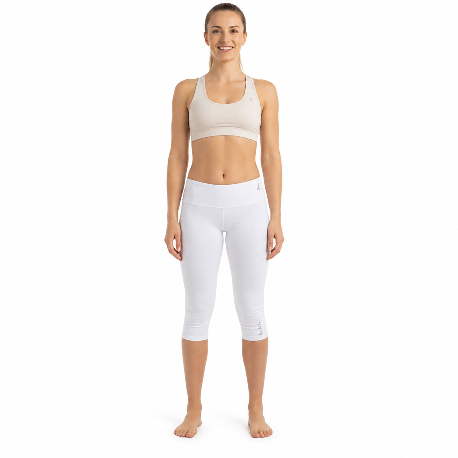 Frontal stehende Person in hellem Sport-BH und weißen Dreiviertel-Leggings vor weißem Hintergrund, barfuß und lächelnd.